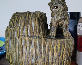 Może przedstawiać: Ceramiczna rzeźba z teksturowaną, oliwkowozieloną podstawą przypominającą pień drzewa z pionowymi żłobieniami. Mała, szczegółowa figurka lwa strażnika z pasującą oliwkowozieloną glazurą stoi po prawej stronie na górnej powierzchni podstawy.