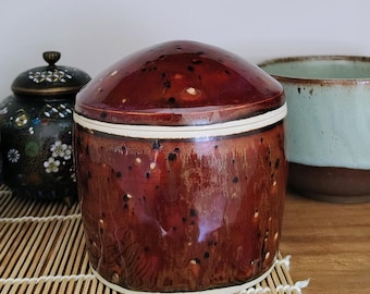 Może przedstawiać: Cylindryczny ceramiczny słoik z błyszczącym, głębokim czerwonobrązowym szkliwem z ciemnobrązowymi i czarnymi plamkami. Słoik posiada dopasowaną, zaokrągloną pokrywkę, która ściśle przylega do góry, tworząc gładki profil z białą krawędzią u podstawy.