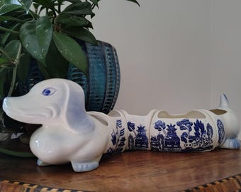 May include: A ceramic dachshund-shaped planter featuring a white base with blue painted details, including a willow pattern design on the body, blue ears, and blue eyes. The elongated dog figure is segmented into three open sections for planting.
