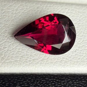 Könnte beinhalten: Ein birnenförmiger, facettierter Edelstein in einer tiefen, leuchtend roten Farbe. Der Stein hat eine spitze Oberseite und eine abgerundete Basis mit mehreren dreieckigen und viereckigen Facetten, die das Licht reflektieren.