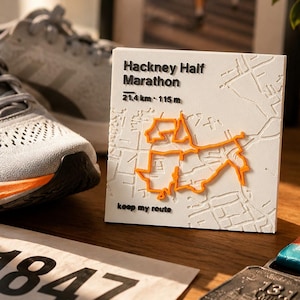 May include: A square white plaque featuring a raised map of the Hackney Half Marathon route in orange. The text reads Hackney Half Marathon, 21.4 km, 115 m, and keep my route. The plaque has a textured surface depicting city streets in relief.