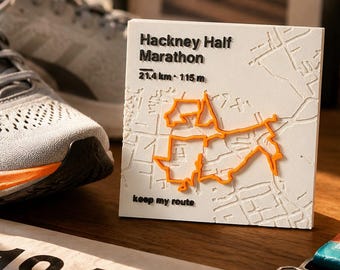 May include: A square white plaque featuring a raised map of the Hackney Half Marathon route in orange, with black text reading Hackney Half Marathon, 21.4 km, 115 m, and keep my route, displayed as a commemorative piece for runners.