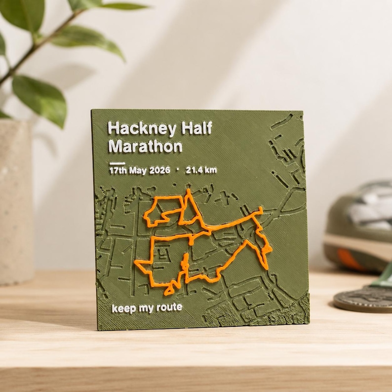 May include: A square olive green plaque featuring a raised orange map route of the Hackney Half Marathon. The text reads Hackney Half Marathon, 17th May 2026, 21.4 km, and keep my route. The surface has a textured finish with embossed details.
