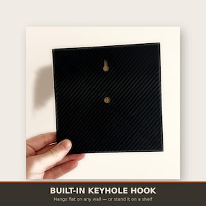 May include: A square black panel featuring a diagonal carbon fibre texture pattern. The back side includes a centred keyhole hook for wall mounting and a small circular indentation below it, designed for versatile display options on a wall or shelf.