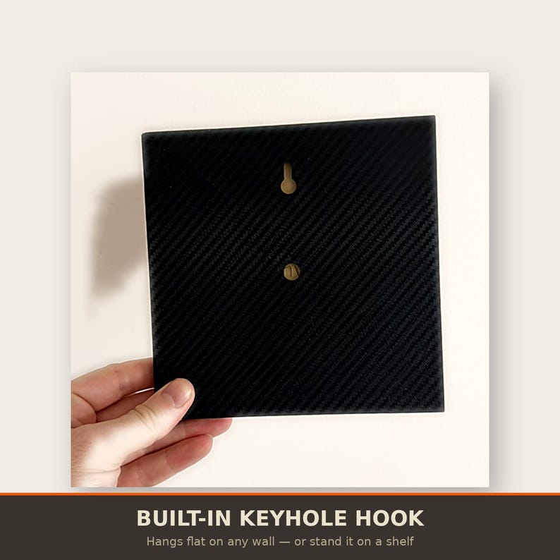 May include: A square black panel featuring a diagonal carbon fibre texture pattern. The back side includes a centred keyhole hook for wall mounting and a small circular indentation below it, designed for versatile display options on a wall or shelf.