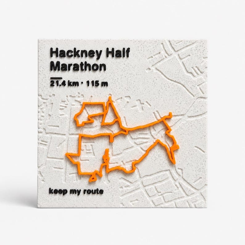 May include: A square white textured plaque featuring a raised orange map outline of the Hackney Half Marathon route. Black text reads Hackney Half Marathon, 21.4 km, 115 m, and keep my route. The background displays a subtle embossed street map.