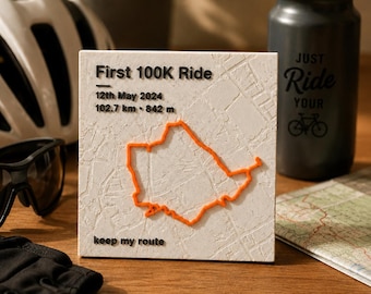 Op de afbeelding: Een vierkante witte plaquette met een verhoogde oranje fietsroutekaart. De tekst luidt: First 100K Ride, 12th May 2024, 102.7 km, 842 m en keep my route. Het oppervlak heeft een gestructureerd kaartpatroon en zwarte letters.