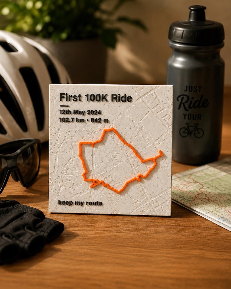 May include: A square white plaque featuring a raised orange cycling route map. Text reads: First 100K Ride, 12th May 2024, 102.7 km, 842 m, and keep my route. The surface has a textured map pattern, and the lettering is in a bold black font.