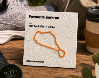 May include: A square white plaque featuring a raised orange map route, black text reading "Favourite parkrun", "12th April 2025", "5.0 km", and "Keep showing up". The plaque displays a textured street map design with embossed lines.