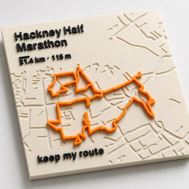 May include: A square off-white plaque featuring a raised orange map route of the Hackney Half Marathon. The text reads Hackney Half Marathon, 21.4 km, 115 m, and keep my route in raised black lettering. The background shows a subtle embossed street map.