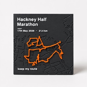 May include: A square black plaque featuring a raised orange map route of the Hackney Half Marathon. The text reads Hackney Half Marathon, 17th May 2026, 21.4 km, and keep my route. The surface displays a textured street grid in relief.