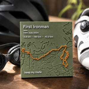 Può includere: Una targa quadrata verde oliva con una mappa in rilievo e una linea di percorso arancione. Il testo recita: First Ironman, 14th July 2024, 3.8 km, 180 km, 42.2 km e keep my route. La mappa mostra i tracciati stradali in rilievo.