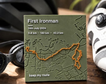 May include: A square olive green plaque featuring a raised map with an orange route line. Text reads: First Ironman, 14th July 2024, 3.8 km, 180 km, 42.2 km, and keep my route. The map displays street layouts in relief, highlighting a specific path.