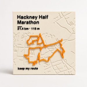 May include: A square off-white plaque featuring a raised orange map route of the Hackney Half Marathon. The text reads Hackney Half Marathon, 21.4 km, 115 m, and keep my route in black. The background displays a subtle embossed street map pattern.