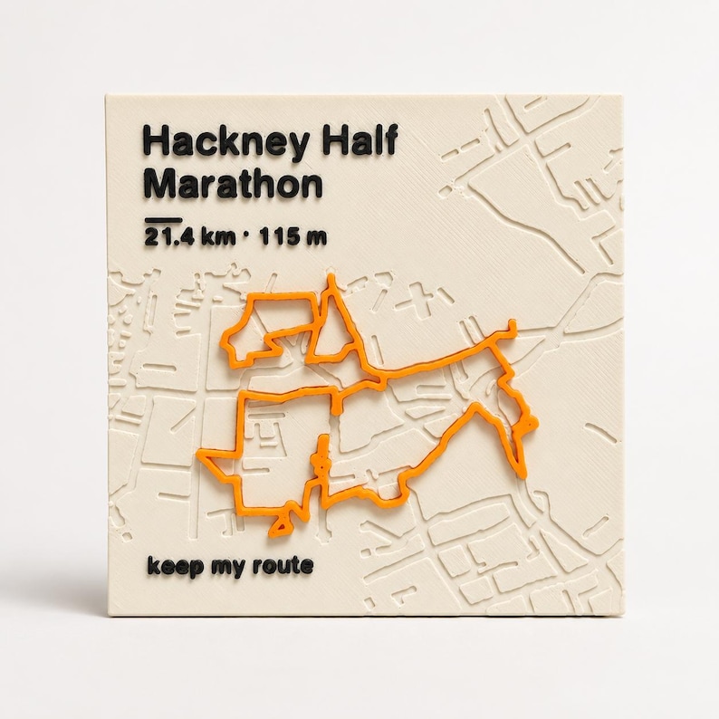 May include: A square off-white plaque featuring a raised orange map route of the Hackney Half Marathon. The text reads Hackney Half Marathon, 21.4 km, 115 m, and keep my route in black. The background displays a subtle embossed street map pattern.
