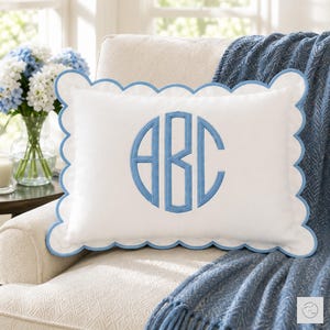 May include: A rectangular white throw pillow featuring a scalloped edge trimmed in light blue piping. The center of the pillow displays a large, circular embroidered monogram with the letters ABC in a matching light blue thread color.