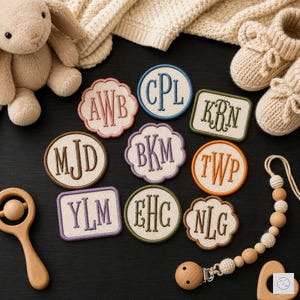 May include: A collection of nine embroidered fabric patches featuring various three-letter monograms in different colors and shapes, including circular, scalloped, and rectangular designs, arranged alongside a plush toy, baby booties, and a wooden rattle.