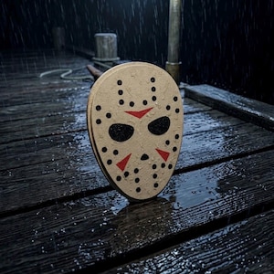 May include: A beige hockey mask featuring a series of black circular ventilation holes, two black eye openings, and three red triangular markings on the forehead and cheeks. The mask is positioned upright on a dark, wet wooden surface during rain.