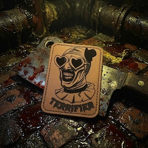 May include: A rectangular tan leather patch featuring a black line art illustration of a clown wearing heart-shaped sunglasses and a ruffled collar, with the text TERRIFIER printed in a bold, dripping font at the bottom of the patch design.