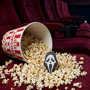 May include: A white and red striped popcorn bucket tipped over on a dark red theater seat, spilling popcorn kernels around a black and white Ghostface mask pin. The bucket features the word POPCORN printed in bold red capital letters.