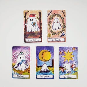 以下が含まれることがあります： ゴーストのイラストが描かれた5枚の長方形のタロットカード。カードには「2 of Sticks」「The World」「Balance」「The Moon」「The Star」と記されています。各カードには、カラフルな手描きの芸術的ディテールとともに、さまざまな設定の白いゴーストキャラクターが描かれています。