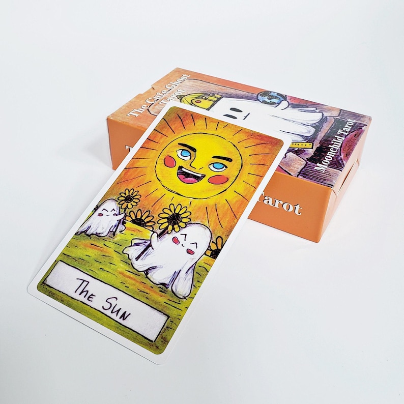 以下が含まれることがあります： 「The Sun」と書かれた長方形のタロットカード。顔のある黄色い太陽、2体の白い幽霊、ヒマワリが描かれています。その背後には「Moonchild Tarot」と記され、幽霊のイラストが描かれたオレンジ色の紙箱が置かれています。