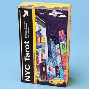 May include: A rectangular deck box for NYC Tarot cards featuring a yellow and black side panel with the text NYC Tarot, Big Apple Divination from the Greatest City on Earth. The front displays a colorful illustration of a busy city street at night.