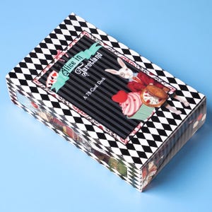 May include: A rectangular tarot card deck box featuring a black and white diamond pattern border. The centre panel displays the text Alice in Tarotland A 78-Card Deck, a white rabbit in a red coat, a pink cupcake, and a pocket watch illustration.