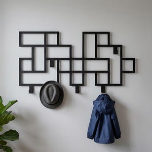 May include: A black wall-mounted coat rack featuring a geometric grid design with multiple rectangular frames and integrated hooks. A grey fedora hat hangs on one hook, and a navy blue hooded jacket hangs on another hook, providing storage.