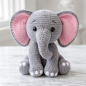 May include: A gray crocheted elephant plush toy with large ears featuring pink inner panels. The elephant has black plastic eyes, a trunk, and white circular accents on the feet. The texture of the yarn is soft and plush, creating a rounded, seated form.