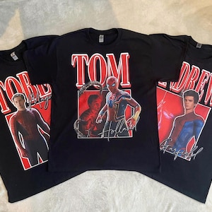 以下が含まれることがあります： スパイダーマン俳優のグラフィックプリントが施された3枚の黒いTシャツ。中央のTシャツには「TOM」「Holland」の文字と共にトム・ホランドが描かれています。左のTシャツには「TOBEY」「Maguire」のトビー・マグワイア、右のTシャツには「ANDREW」「Garfield」のアンドリュー・ガーフィールドが描かれています。