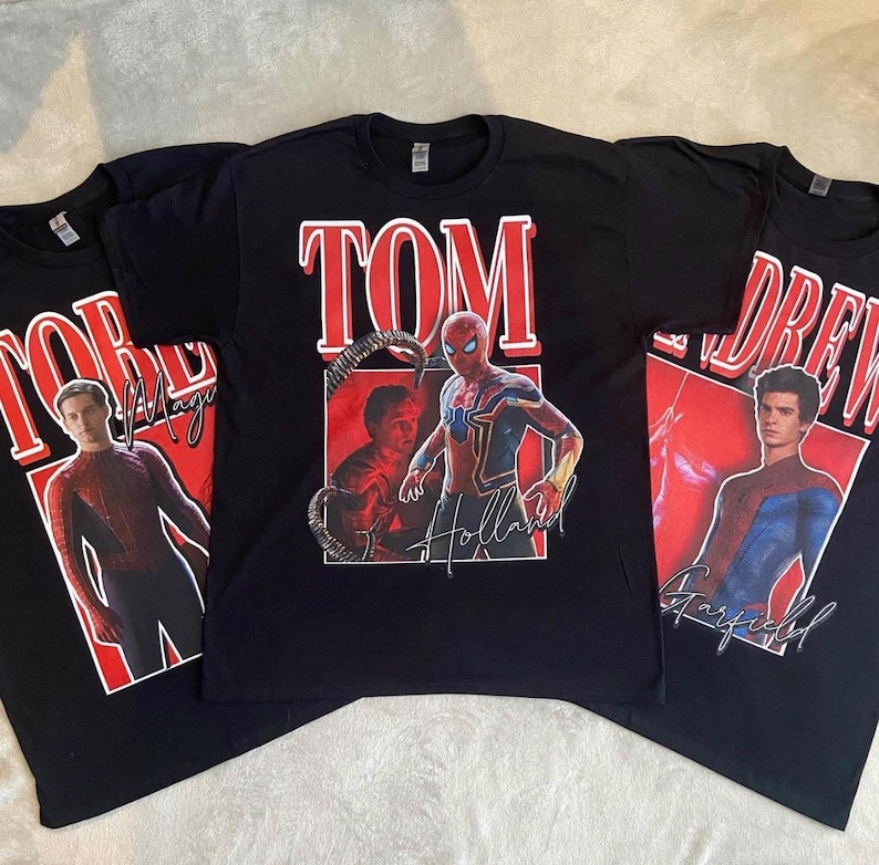 以下が含まれることがあります： スパイダーマン俳優のグラフィックプリントが施された3枚の黒いTシャツ。中央のTシャツには「TOM」「Holland」の文字と共にトム・ホランドが描かれています。左のTシャツには「TOBEY」「Maguire」のトビー・マグワイア、右のTシャツには「ANDREW」「Garfield」のアンドリュー・ガーフィールドが描かれています。