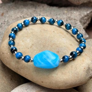 May include: A beaded bracelet featuring a series of round, dark blue tiger eye beads alternating with small, polished silver-tone metal spacer beads, centred with a single, larger, smooth, irregular-shaped bright blue glass bead.