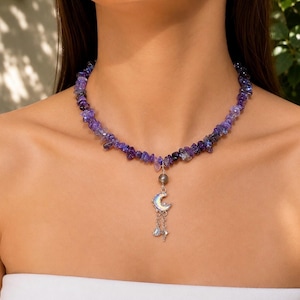 May include: A choker necklace featuring irregular, tumbled purple amethyst-coloured stone chips strung together. A silver-toned pendant hangs from the centre, consisting of a small grey bead, a crescent moon charm, and two delicate dangling chains.