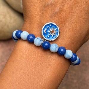 Peut inclure: Un bracelet de perles alternant des perles bleu foncé et bleu clair marbrées. Une breloque circulaire argentée est suspendue au bracelet, présentant un fond en émail bleu avec un motif de soleil et de croissant de lune argenté, orné d'une petite pierre orange.