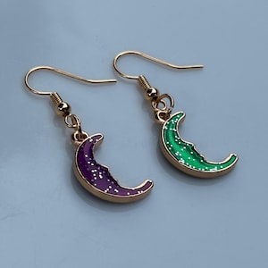 Può includere: Un paio di orecchini pendenti con ciondoli a forma di mezzaluna con bordi in metallo dorato. Un ciondolo è viola con accenti di glitter bianchi, l'altro è verde con accenti di glitter bianchi. Entrambi pendono da ganci in metallo dorato.