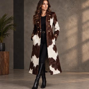 May include: A long, double-breasted coat featuring a brown and white cow-print pattern, gold-toned buttons, and structured lapels. The coat is worn over a black top, black trousers with a belt, and black knee-high boots, creating a full-length look.