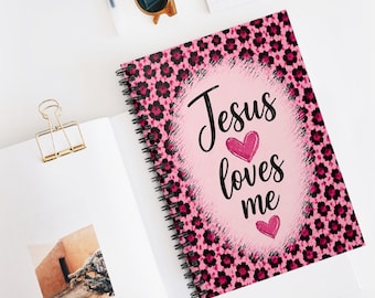May include: A spiral-bound notebook featuring a pink and black leopard print pattern. The cover displays a light pink oval center with the black text "Jesus loves me" and two pink hearts. A gold metal binder clip is attached to the open page.