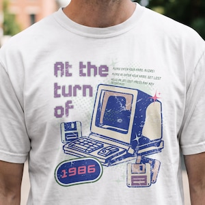 Peut inclure: Un t-shirt blanc à manches courtes avec un motif représentant un ordinateur vintage, deux disquettes et le texte "At the turn of 1986". Le graphique inclut de petits textes : "PLEASE ENTER YOUR NAME: ADORE!", "PLEASE RE-ENTER YOUR NAME: GET LOST" et "HELLO MR. GET LOST! PRESS ANY KEY TO CONTINUE!"