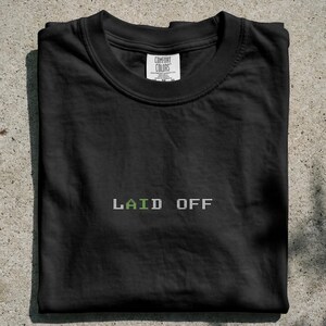 May include: A black crew neck t-shirt featuring the text LAID OFF embroidered in a pixelated font style. The letters are primarily white with a single green letter A. The shirt includes a Comfort Colors brand label at the inner neckline.