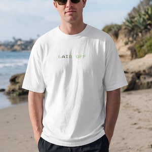 May include: A white short-sleeved t-shirt featuring the text "LAID OFF" printed in a pixelated, light green font across the center of the chest. The garment has a classic crew neckline and a relaxed fit, constructed from a solid white fabric.