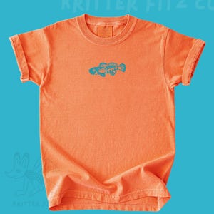 May include: A short-sleeved orange t-shirt featuring a teal graphic of a fish skeleton centered on the chest. The text KRITTER FITZ CO is printed in light blue at the top, and a small light blue animal logo is visible in the bottom left corner.