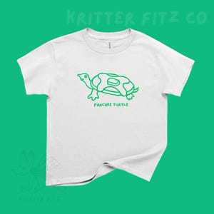 May include: A white short-sleeve t-shirt featuring a green line-art graphic of a turtle with a pancake on its back. Below the turtle, the text reads PANCAKE TURTLE in green capital letters. The shirt has a crew neckline and a standard fit.