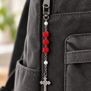 May include: A silver-toned metal zipper pull charm featuring three small red rose beads, two white pearl-like beads, and a textured silver cross pendant hanging from a dark gray canvas backpack zipper.
