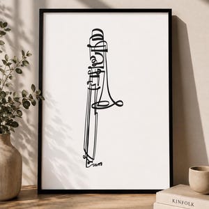 Puede incluir: Una lámina de arte enmarcada que presenta un dibujo de línea negra de un trombón sobre un fondo blanco. La ilustración utiliza un trazo continuo y fluido para formar la silueta del instrumento musical, dentro de un marco negro sencillo.