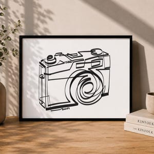 Może przedstawiać: Czarno-biały wydruk grafiki liniowej przedstawiającej aparat fotograficzny w cienkiej czarnej ramce. Dzieło składa się z ciągłej czarnej linii tuszu tworzącej kształt aparatu w stylu vintage na białym tle, w orientacji poziomej prostokątnej.