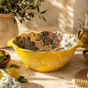 Può includere: Una ciotola in ceramica giallo brillante con bordo smerlato e due manici laterali. L'esterno presenta un motivo floreale bianco, mentre l'interno è decorato con un intricato design in stile mandala nei toni del rosso, blu e verde.