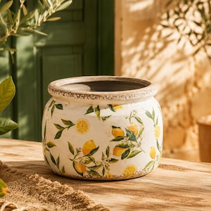 Op de afbeelding: Een ronde keramische plantenpot met een witte basis, versierd met een patroon van gele citroenen, groene bladeren en kleine witte bloesems. De rand heeft een verweerde, textuurrijke bruine afwerking en de binnenkant is donkergrijs.