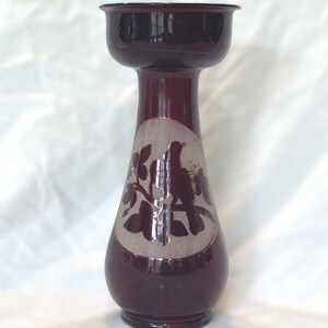 May include: A deep purple glass vase with a flared rim and a tapered neck. The body features a circular frosted panel containing a dark purple silhouette of a bird perched on a leafy branch, contrasting against the translucent glass surface.