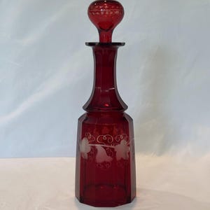 May include: A tall, deep red glass decanter with a matching spherical stopper. The decanter features a faceted hexagonal base and a slender neck. A delicate, etched white floral vine pattern wraps around the midsection of the glass body.